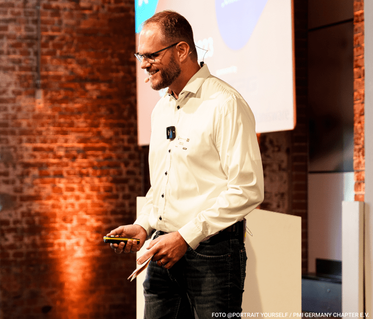 Patric Eid präsentiert seitlich stehend mit einem Präsentationsklicker in der Hand. Im Hintergrund ist eine PowerPoint-Folie bi der Eröffnung des PM-Summits 2023 zu sehen.